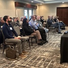 Engaged audience at a professional seminar in a well-lit conference room with a speaker.