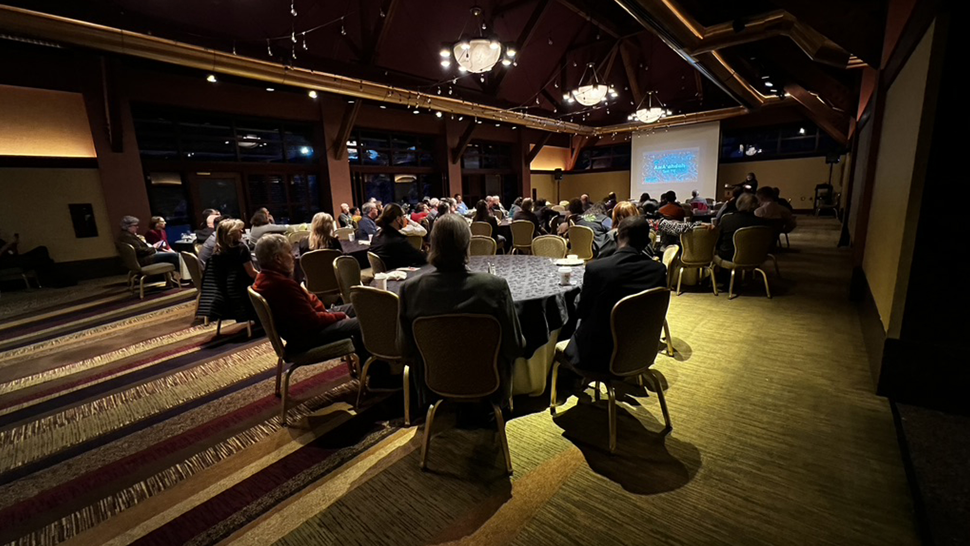 Diverse attendees engage in a warm, inviting indoor event with presentations and elegant decor.