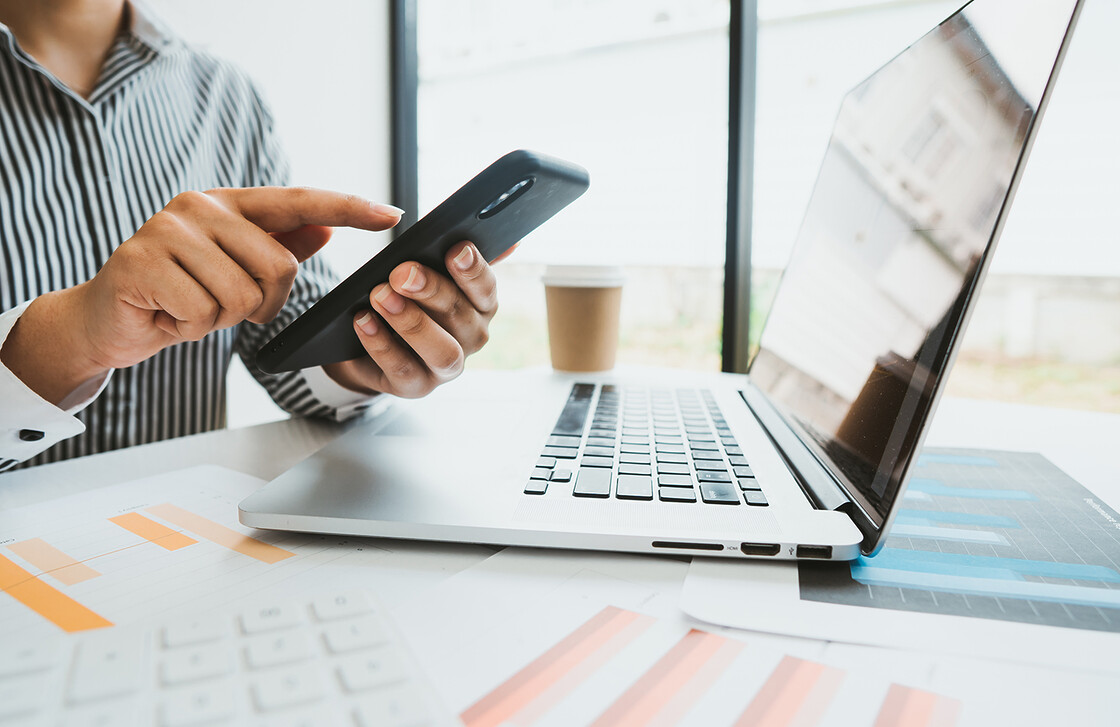 Professional multitasking in a bright modern workspace with laptop, smartphone, and coffee cup.